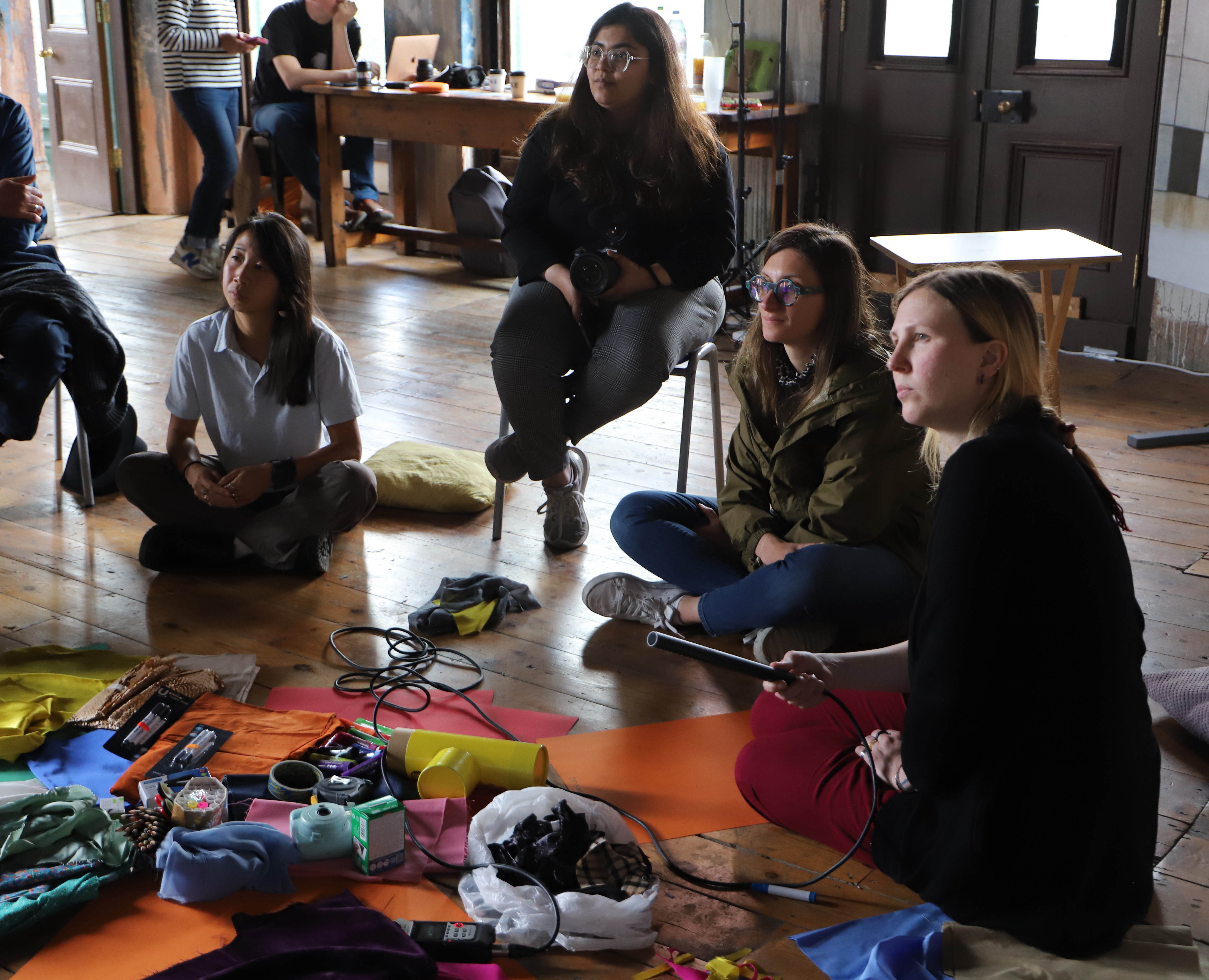 Chiara and Abbey sitting on the floor of the Old Waiting Room in Peckham, together with the workshop participants. On the floor in front of them crafts material.