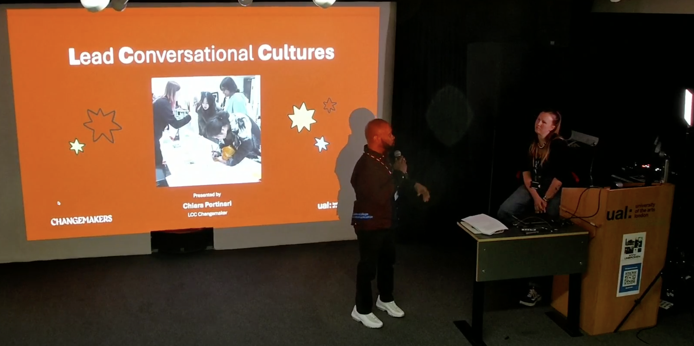 Kevin J Brazant introduces Chiara Portinari's presentation titled Lead Conversational Cultures, visible on a screen behind his back. Chiara is sitting on the side waiting to start their presentation.